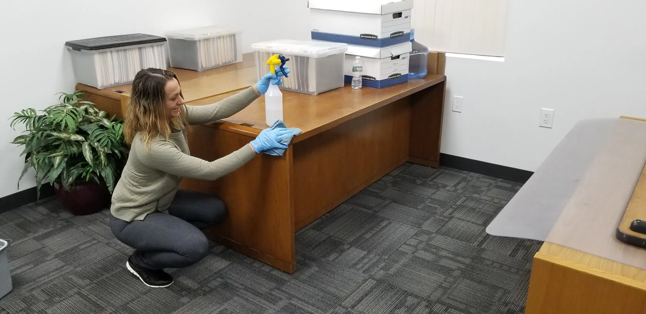 Crew member in gloves wiping down a wooden office desk with a spray bottle and cloth during office cleaning